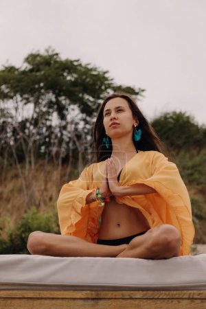 Photo for Confident young woman in boho styled crop top sitting in lotus position while meditating seaside - Royalty Free Image