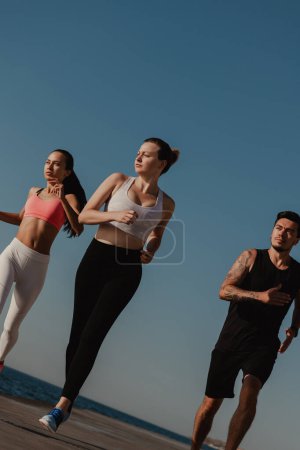 Téléchargez les photos : Trois jeunes gens confiants en vêtements de sport semblent concentrés tout en faisant du jogging au bord de la mer ensemble - en image libre de droit
