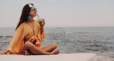 Photo for Beautiful young woman in boho styled crop top drinking cocktail and smiling while relaxing seaside - Royalty Free Image
