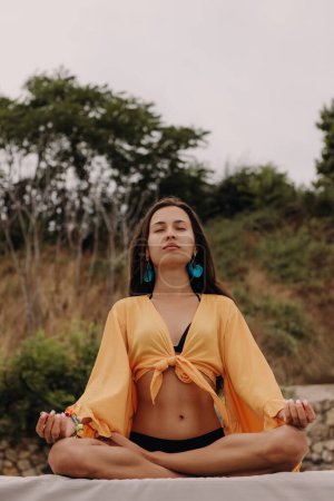 Photo for Beautiful young woman in boho styled crop top sitting in lotus position while meditating seaside - Royalty Free Image