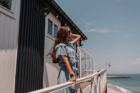 Téléchargez les photos : Belle jeune femme en robe d'été ajustant les cheveux tout en se relaxant sur la terrasse vue mer - en image libre de droit