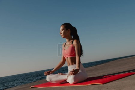 Foto de Atractiva joven en ropa deportiva meditando mientras está sentada en posición de loto junto al mar - Imagen libre de derechos