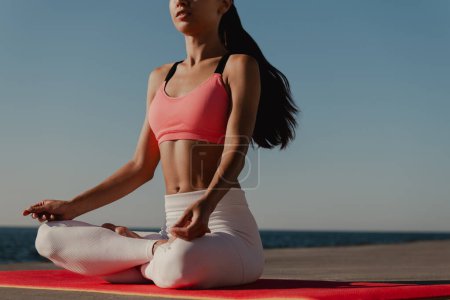 Foto de Primer plano de mujer irreconocible en ropa deportiva meditando mientras está sentada en posición de loto junto al mar - Imagen libre de derechos