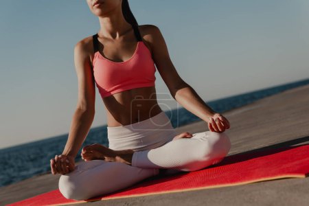 Foto de Primer plano de una joven irreconocible en ropa deportiva meditando mientras está sentada en posición de loto junto al mar - Imagen libre de derechos