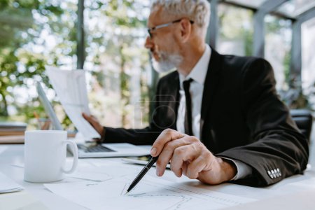 Téléchargez les photos : Homme d'affaires mature confiant pointant le papier avec stylo tout en analysant les graphiques et les graphiques dans le bureau - en image libre de droit