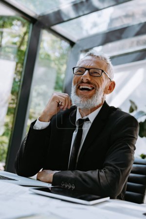 Téléchargez les photos : Homme d'affaires mature confiant souriant tout en étant assis à son lieu de travail dans le bureau moderne - en image libre de droit
