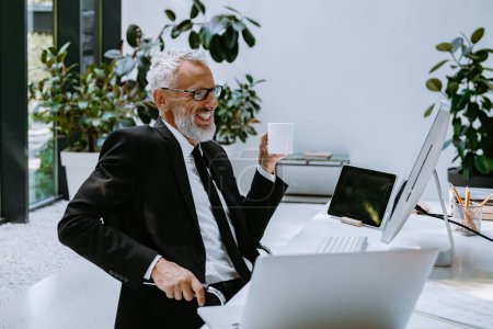 Téléchargez les photos : Heureux homme mature en coffret tenant tasse de café tout en travaillant dans le bureau moderne - en image libre de droit