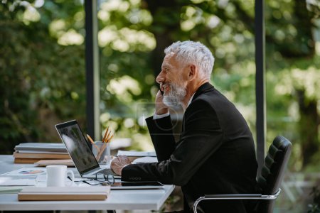Téléchargez les photos : Homme d'affaires mature confiant parlant sur un téléphone portable assis son lieu de travail dans le bureau - en image libre de droit