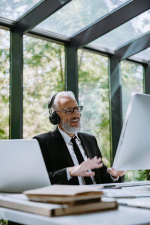 Foto de Confiado hombre de negocios maduro en los auriculares usando la computadora mientras que tiene conferencia de la tela de oficina - Imagen libre de derechos