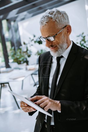 Téléchargez les photos : Homme d'affaires mature concentré utilisant une tablette numérique tout en restant debout dans le bureau - en image libre de droit