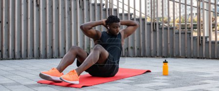 Téléchargez les photos : Beau jeune homme africain en vêtements de sport faire des sit-ups tout en faisant de l'exercice à l'extérieur - en image libre de droit