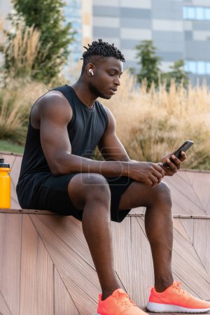 Téléchargez les photos : Homme africain confiant dans les vêtements de sport vérifier son programme de remise en forme tout en utilisant un téléphone intelligent à l'extérieur - en image libre de droit