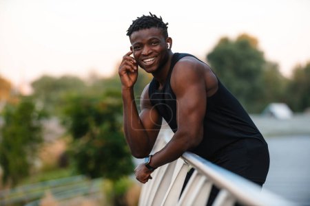 Téléchargez les photos : Homme africain joyeux en vêtements de sport ajuster ses écouteurs tout en se relaxant après l'entraînement en plein air - en image libre de droit