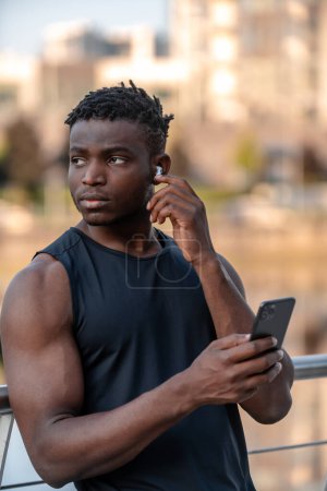 Foto de Joven deportista africano ajustando auriculares y sosteniendo el teléfono inteligente mientras descansa después de entrenar al aire libre - Imagen libre de derechos