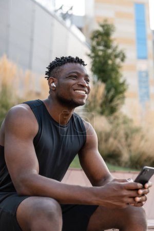 Téléchargez les photos : Heureux homme africain en vêtements de sport en utilisant un téléphone intelligent tout en faisant une pause après l'entraînement en plein air - en image libre de droit