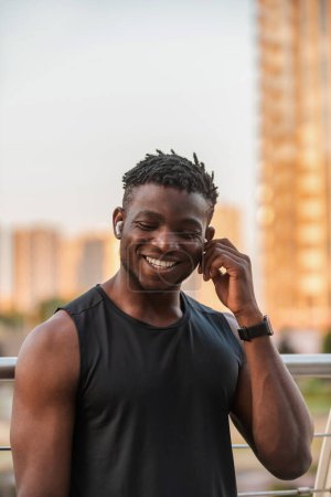 Téléchargez les photos : Heureux homme africain en vêtements de sport ajuster ses écouteurs tout en se reposant après l'entraînement à l'extérieur - en image libre de droit