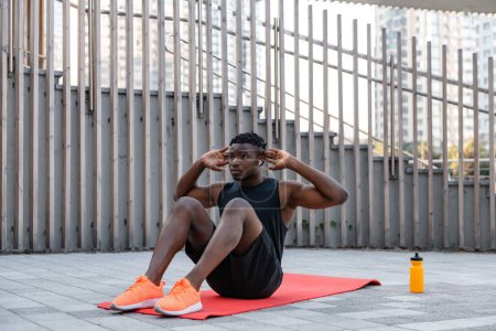 Téléchargez les photos : Confiant jeune homme africain en vêtements de sport faire des sit-ups tout en faisant de l'exercice à l'extérieur - en image libre de droit
