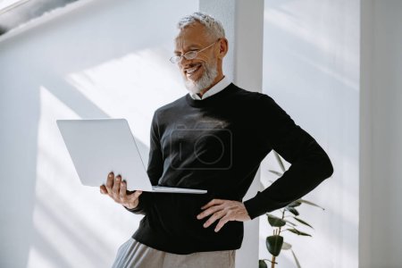 Téléchargez les photos : Heureux homme mature en tenue décontractée intelligente regardant ordinateur portable tout en se tenant dans le bureau - en image libre de droit