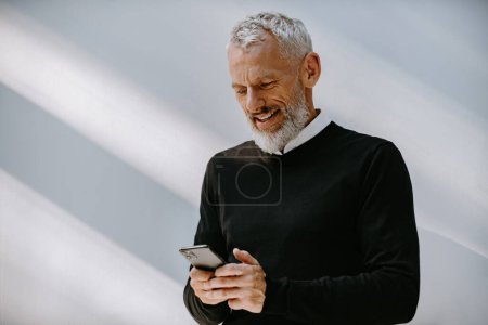 Téléchargez les photos : Portrait d'un homme âgé confiant utilisant un téléphone intelligent et souriant tout en se tenant contre le mur - en image libre de droit