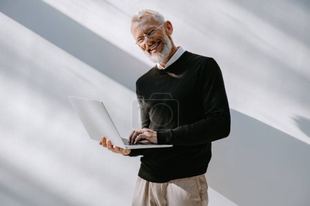 Téléchargez les photos : Homme mature confiant en tenue décontractée intelligente portant un ordinateur portable et souriant tout en se tenant contre un mur - en image libre de droit