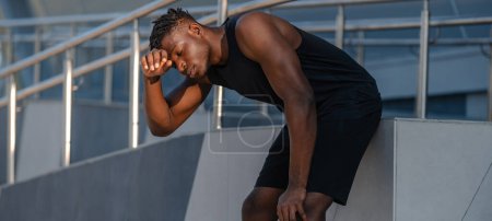 Téléchargez les photos : Homme africain épuisé en vêtements de sport se penchant les mains sur les genoux tout en se reposant après l'entraînement à l'extérieur - en image libre de droit