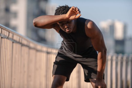 Téléchargez les photos : Jeune athlète africain couvrant le visage avec la main et regardant fatigué après l'entraînement sportif à l'extérieur - en image libre de droit
