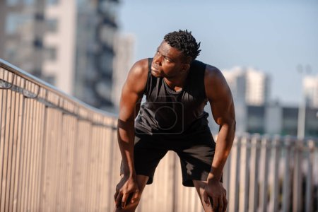 Téléchargez les photos : Fatigué athlète africain penché les mains sur les genoux tout en se reposant après l'entraînement à l'extérieur - en image libre de droit