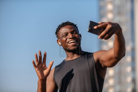 Foto de Feliz joven atleta africano utilizando el teléfono inteligente y sonriendo mientras tiene videollamada al aire libre - Imagen libre de derechos
