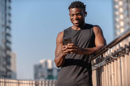 Téléchargez les photos : Athlète africain heureux utilisant un téléphone intelligent tout en vérifiant son programme de remise en forme avant de s'entraîner à l'extérieur - en image libre de droit