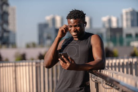 Téléchargez les photos : Homme africain joyeux en vêtements de sport en utilisant un téléphone intelligent et en ajustant les écouteurs tout en se reposant après l'entraînement - en image libre de droit