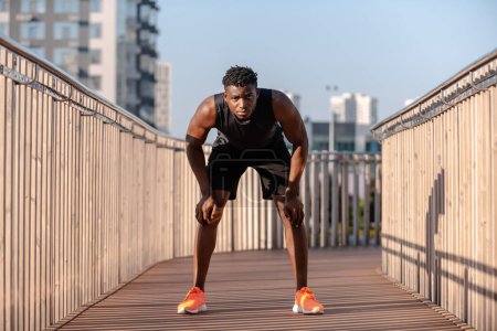Téléchargez les photos : Athlète africain fatigué en vêtements de sport se penchant les mains sur les genoux tout en se reposant après l'entraînement à l'extérieur - en image libre de droit