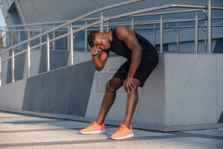 Téléchargez les photos : Toute la longueur de l'homme africain fatigué en vêtements de sport se reposant après l'entraînement sportif à l'extérieur - en image libre de droit