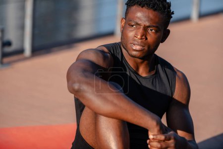 Téléchargez les photos : Confiant jeune homme africain en vêtements de sport se reposant après l'entraînement tout en étant assis sur tapis d'exercice à l'extérieur - en image libre de droit