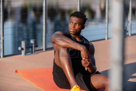 Téléchargez les photos : Beau jeune homme africain en vêtements de sport se reposant après l'entraînement tout en étant assis sur tapis d'exercice à l'extérieur - en image libre de droit