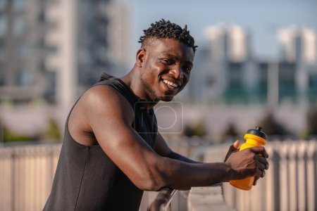 Téléchargez les photos : Heureux homme africain en vêtements de sport tenant bouteille d'eau tout en se reposant après l'entraînement à l'extérieur - en image libre de droit