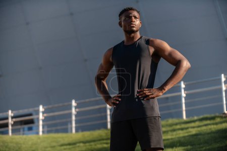 Téléchargez les photos : Homme africain confiant en vêtements de sport tenant la main sur la hanche pendant l'entraînement à l'extérieur - en image libre de droit