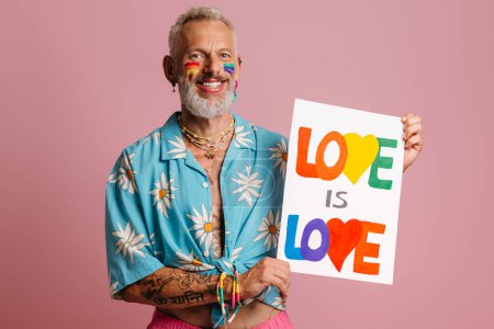 Foto de Feliz maduro gay hombre con arco iris bandera maquillaje mostrando colorido banner contra rosa fondo - Imagen libre de derechos