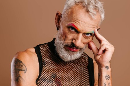 Téléchargez les photos : Portrait d'homme gay mature barbu avec des sourcils colorés arc-en-ciel debout sur fond brun - en image libre de droit
