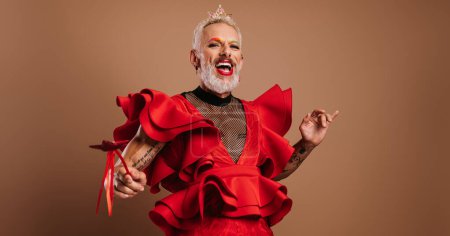 Téléchargez les photos : Homme gay ludique avec couronne sur la tête portant belle robe rouge sur fond brun - en image libre de droit