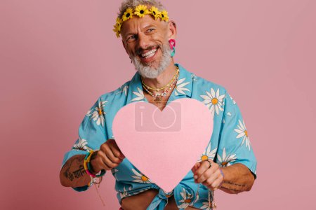 Foto de Feliz maduro gay hombre en floral guirnalda celebración corazón forma y sonriendo contra rosa fondo - Imagen libre de derechos