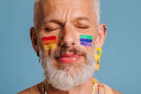 Téléchargez les photos : Portrait d'homme gay mature avec drapeau arc-en-ciel maquillage gardant les yeux fermés sur fond bleu - en image libre de droit