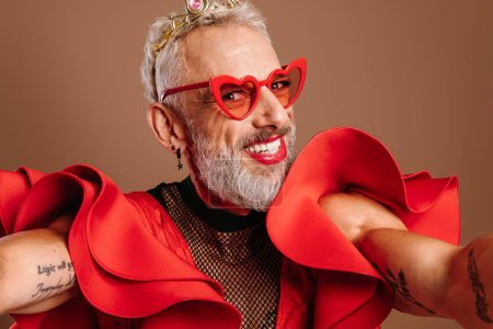 Foto de Feliz maduro gay hombre en hermosa rojo vestido haciendo auto retrato contra marrón fondo - Imagen libre de derechos