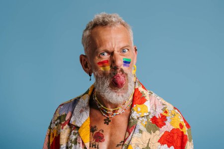 Foto de Pensativo maduro gay hombre con arco iris bandera maquillaje celebración de la mano en la barbilla contra fondo azul - Imagen libre de derechos