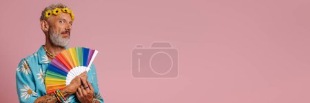 Téléchargez les photos : À la mode mature gay homme dans floral couronne agitant ventilateur de main tout en se tenant debout sur fond rose - en image libre de droit