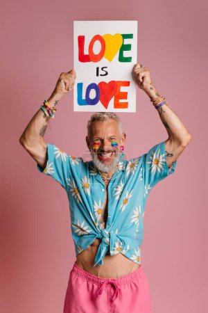 Téléchargez les photos : Heureux homme gay mature avec drapeau arc-en-ciel maquillage tenant bannière sur la tête sur fond rose - en image libre de droit