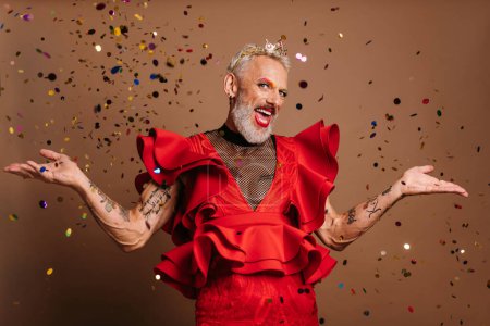 Foto de Alegre gay hombre en hermosa rojo vestido gesto mientras confeti volando alrededor de él contra marrón fondo - Imagen libre de derechos