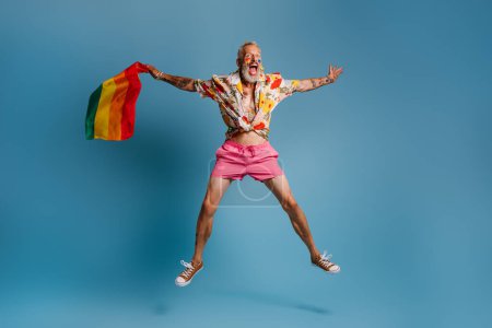 Foto de Longitud completa de maduro gay hombre llevando arco iris bandera y sonriendo mientras saltar contra azul fondo - Imagen libre de derechos