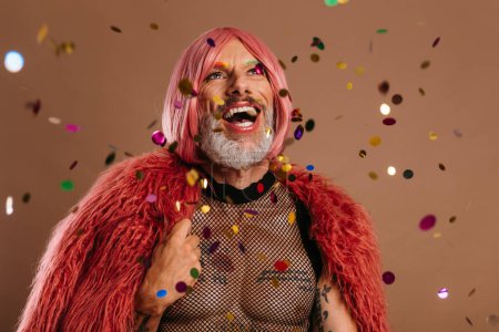 Foto de Feliz gay hombre en rosa peluca mirando el colorido confeti volando alrededor contra marrón fondo - Imagen libre de derechos
