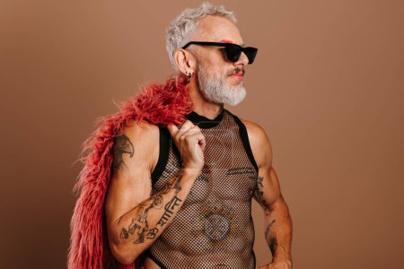 Téléchargez les photos : Homme gay mature à la mode en chemise transparente portant manteau moelleux sur l'épaule sur fond brun - en image libre de droit