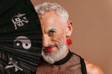 Foto de Barbudo maduro gay hombre con color cejas cubriendo medio cara con ventilador contra marrón fondo - Imagen libre de derechos